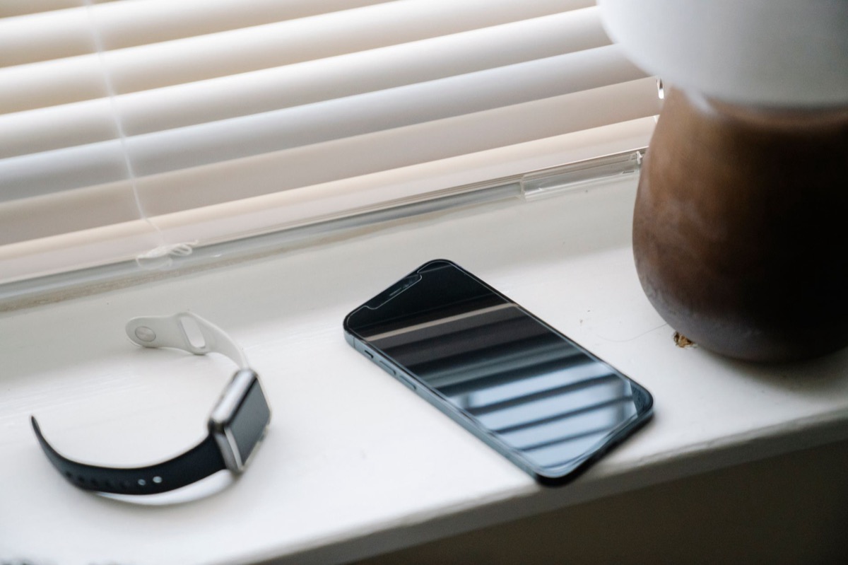 A phone resting on a sunlit windowsill beside a watch, soft daylight coming through half-open blinds