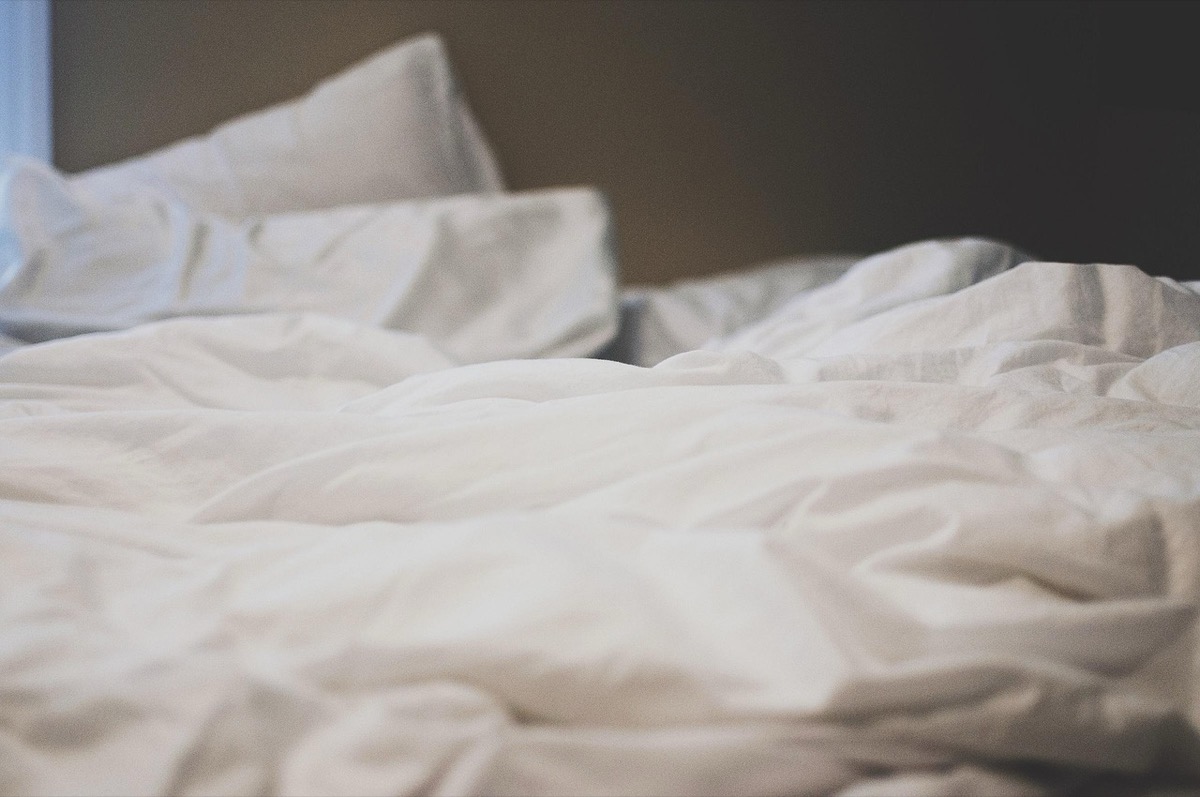 An empty, unmade bed in soft morning light, the pillows still holding the shape of someone who has gone